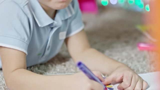 Tilt down of blonde little boy lying on carpet and writing on handmade greeting card with felt tip pen