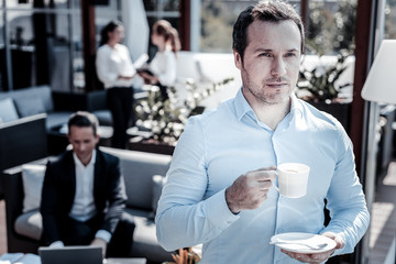 Favorite espresso. Serious nice self employed man holding a coffee cup and looking in front of him while standing in the cafe