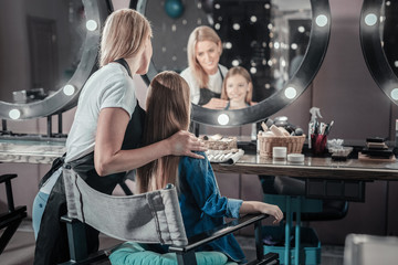 Look at yourself. Pleasant skillful makeup artist standing behind her client and smiling while...