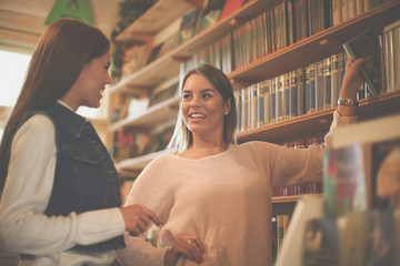 Two young students girl taking book in library and having conversation.