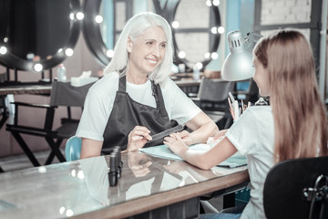 Neat manicure. Positive delighted experienced woman smiling and filing her clients nails while working in the beauty salon