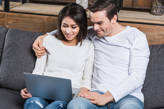 smiling multiethnic couple using laptop together while sitting on sofa at home