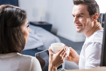 Fototapeta premium multiethnic couple holding cups of coffee and smiling each other at home