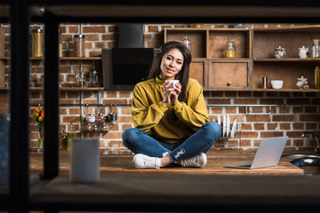 Fototapeta na wymiar beautiful young african american woman holding cup of coffee and smiling at camera while sitting on wooden kitchen table with laptop