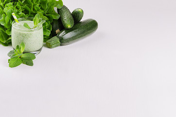 Green vegetable smoothie in glass with deep green vegetables and straw, mint, copy space. Soft white wooden board background.