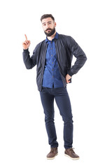 Smart casual bearded hipster in bomber jacket pointing finger up looking at camera. Full body length isolated on white background. 