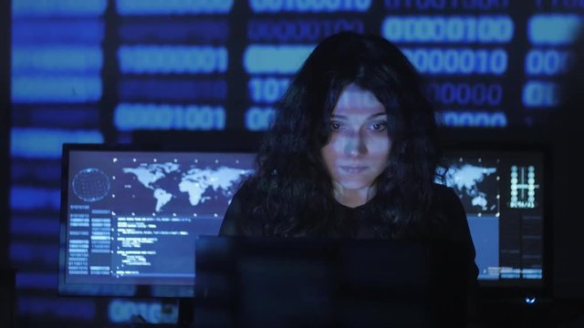 Woman Hacker programmer with curly hair is working on computer in cyber security center filled with display screens. Binary code on her face