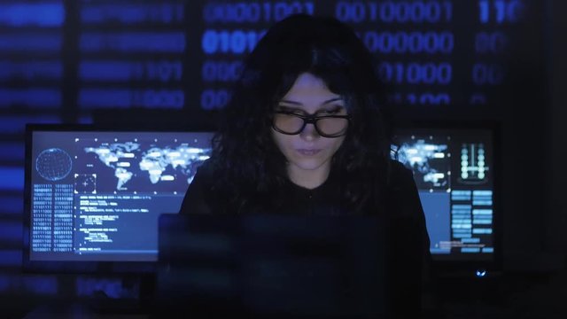 bespectacled Woman Hacker programmer is working on computer in cyber security center filled with display screens. Binary code on her face