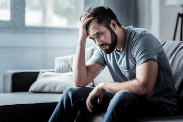 Unpleasant pain. Sad unhappy handsome man sitting on the sofa and holding his forehead while having headache - obrazy, fototapety, plakaty