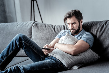 Boring TV shows. Unhappy depressed bored man hugging a cushion and holding a remote control while...