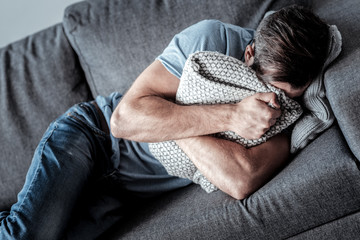 Unhappy life. Cheerless sad depressed man lying on the sofa and holding a cushion while crying in it