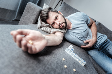 Suicide attempt. Cheerless unhappy young man lying on the sofa and holding pills while trying to...