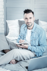 Fototapeta premium Positive mood. Delighted handsome positive man holding a notebook and looking at you while sitting on the bed