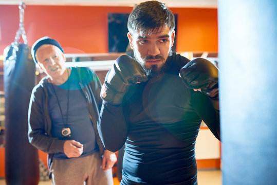 Waist up portrait of bearded Middle-Eastern fighter hitting punching bag at boxing practice in martial arts club with senior coach in background