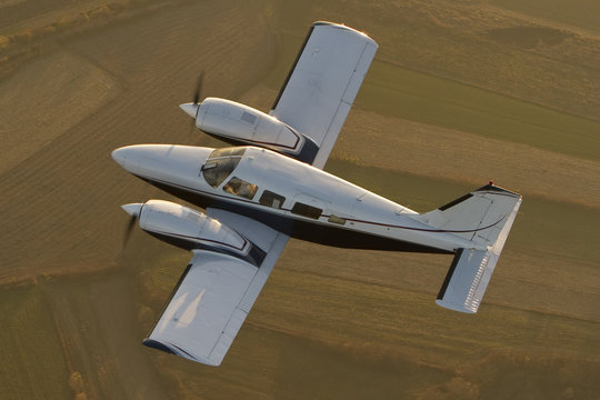 Realistic image of white two engine private plane flying over the farm fields during the warm sunset 