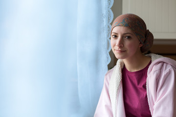 Young positive adult female cancer patient sitting in the kitchen by a window, smiling and looking...