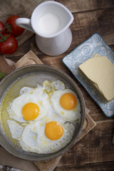 Fried eggs in a frying pan with tomatoes, milk and butter for breakfast on a wooden background.