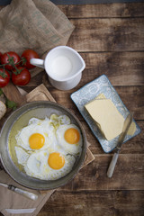 Fried eggs in a frying pan with tomatoes, milk and butter for breakfast on a wooden background.