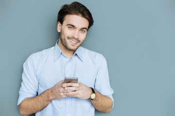 Modern igen. Happy nice handsome man holding his smartphone and smiling to you while typing a message