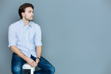 I am tired. Pleasant sad young man sitting on a stool and holding his hands together while thinking...