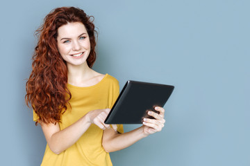 Digital technology. Attractive positive young woman smiling and looking at you while using a modern tablet
