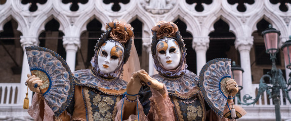 Two women in traditional costume and painted masks, with decorated fans, standing in front ofthe...