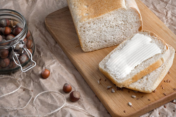 A loaf of wheat white bread, with a cut piece, lies on a wooden table and packing kraft paper. Next to the nuts in a glass jar.