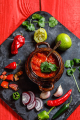 Homemade salsa with ingredients on black stone desk on red rustic background, tomato, pepper, onion, chili, habanero, coriander leaf. Mexican food concept, top view, overhead