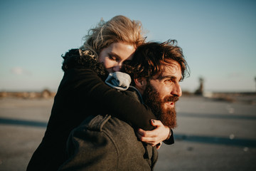 Cool indie couple having fun outdoors while he gives her a piggyback.