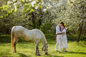 young couple in national Ukrainian clothes with white horse in a flowery garden