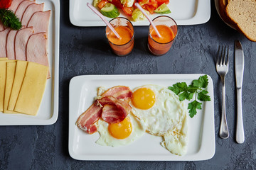 English Breakfast with scrambled eggs and bacon, carrot juice, bread, sliced cheese and ham, on a dark stone background