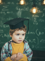 Smart child in graduate cap on serious face, shy, holds hands crossed. Kid, preschooler or first former, chalkboard on background, defocused. Graduate concept. Boy looks cute in square academic cap.