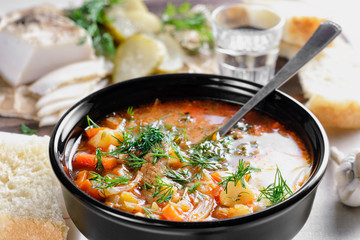 Appetizing Russian borscht in a black ceramic bowl is on the table next to sliced lard and pickled cucumber, garlic, slices of bread and a glass of vodka on a wooden plank