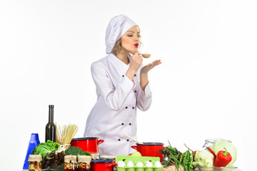 Female chef wearing white uniform holds wooden spoon with hot food. Plump woman cooking soup with vegetables on kitchen. Professional cook in white suit standing at table. Cooking dinner - tasting.