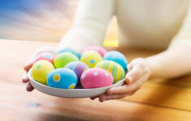 Fototapeta na wymiar easter, religious holidays and people concept - close up of woman hands with colored eggs on plate over sky background