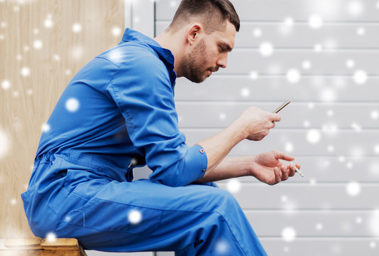 bad habits and people concept - auto mechanic with smartphone smoking cigarette at workshop over snow