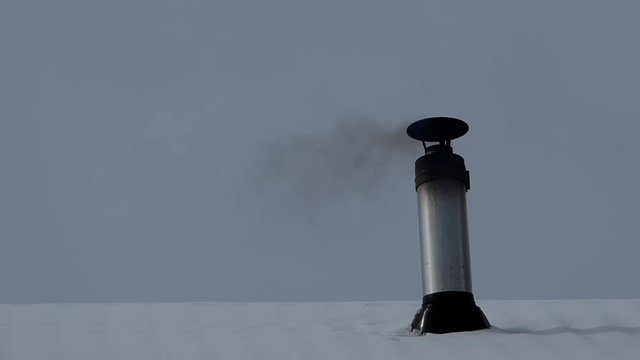 Gray iron stovepipe with lot of soot, winter sky behind. Black smoke flying away. House roof with snow in the village.