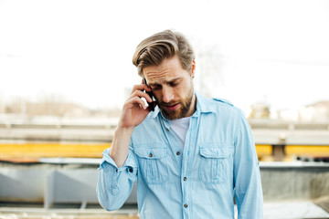 casual young man in blue shirt having a conversation, talking on cellphone, somewhere outside, smiling and enjoying this conversation