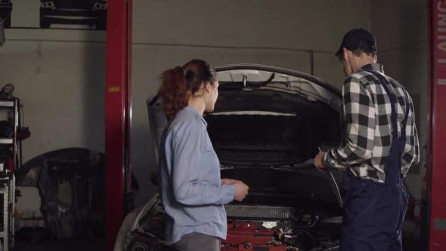 Cute car or automotive technician wearing work robe and a cap writes down in his notepad complaints from his young female client, looking at her vehicle at the same time.
