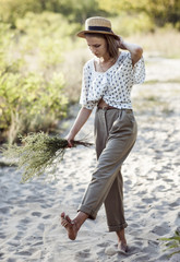 stylish teenager girl in rolled light pants and in a straw hat walks barefoot in the sand in the...