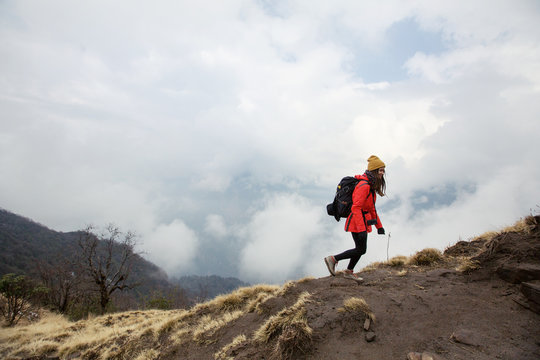 Young traveller woman wearing backpack, red jacket and yellow hat walking  in fog mountain trekking above clouds and valley