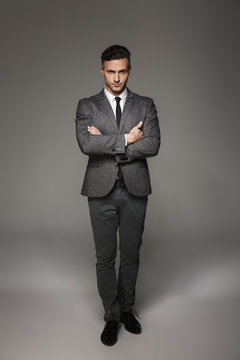 Full length portrait of masculine man wearing business suit posing on camera with serious look keeping arms folded, isolated over gray background