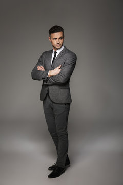 Full length portrait of fashionable man wearing business suit looking aside with focused gaze keeping arms crossed, isolated over gray background