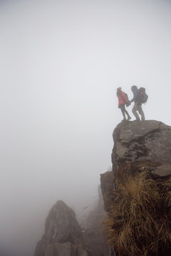 Hikers couple with backpacks relaxing on top of  mountain cliff in white fog, enjoying view of valley above clouds
