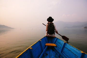 Woman wearing black hat and backpack sitting on boat with paddle, nice mountain lake Fewa and sunset on background