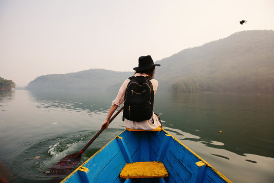 Traveller female wearing black hat and backpack sitting on boat with paddle, nice mountain lake Fewa on background