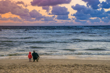 Obraz premium Father and little child watching the sunset on Baltic Sea with cloudy beautiful sky and wavy water