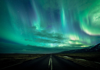 Night scene of a road leading towards distance with Northern lights aka Aurora Borealis glowing on the sky with mountains in Iceland