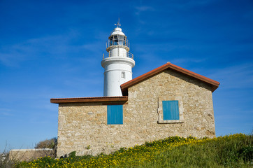 Lighthouse, white lighthouse on blue sky background. Summer, spring, sea and hope concept. Stone building with red roof tiles and blue windows. Green field with yellow flowers. Сyprus, Paphos.