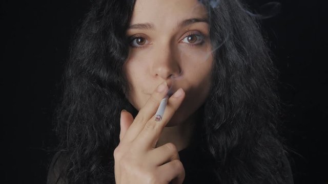 Portrait of young brunette woman with curly hair is smoking a cigarette and looking at the camera on the dark background